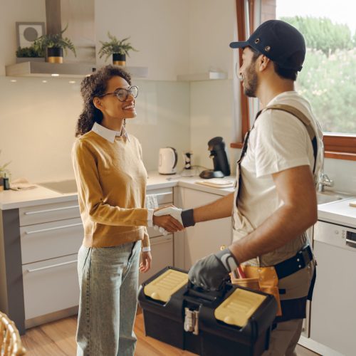 Professional male handyman shaking hands with happy female client while standing at home kitchen